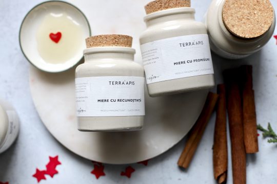 Borcan ceramic Apiarium Miere cu Recunoștință și Miere cu Promisiune, așezate pe un platou alb, alături de miere cu inimă roșie și batoane de scorțișoară, decor festiv de iarnă.”