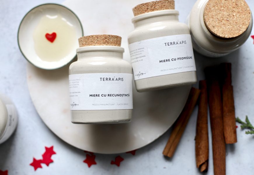 Borcan ceramic Apiarium Miere cu Recunoștință și Miere cu Promisiune, așezate pe un platou alb, alături de miere cu inimă roșie și batoane de scorțișoară, decor festiv de iarnă.”